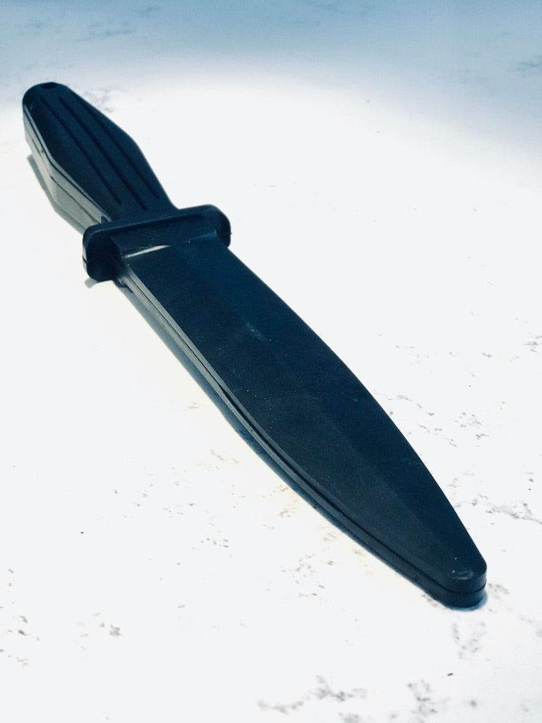 Full-length black rubber training knife lying diagonally on a light surface, showing its rounded tip and grooved handle.